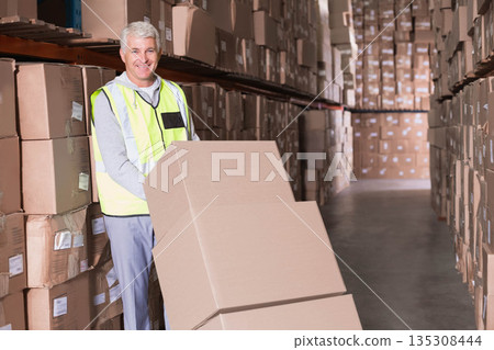 Senior male warehouse worker wearing neon vest pushing dolly with cardboard boxes down rack aisle 135308444