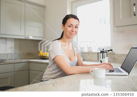 Adult woman typing on laptop at marble island with mug, French press, fruit bowl, gray cabinets 135308450