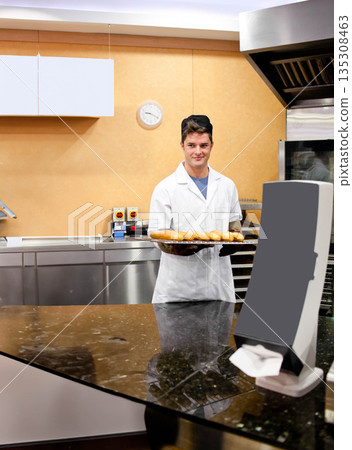 Adult male baker wearing chef jacket standing behind counter in bakery holding tray of bread rolls Adult male baker wearing chef jacket standing behind counter in bakery holding tray of bread rolls 135308463