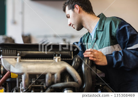 Man wearing two-tone coverall leaning over engine inspecting manifold hose in garage, copy space Man wearing two-tone coverall leaning over engine inspecting manifold hose in garage, copy space 135308469