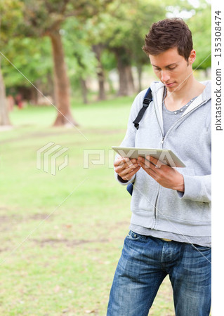 Man standing on grassy lawn in park using tablet and carrying backpack, copy space 135308474