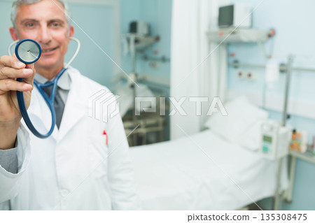 Senior doctor offering stethoscope to viewer in hospital room with bed, monitor, copy space 135308475