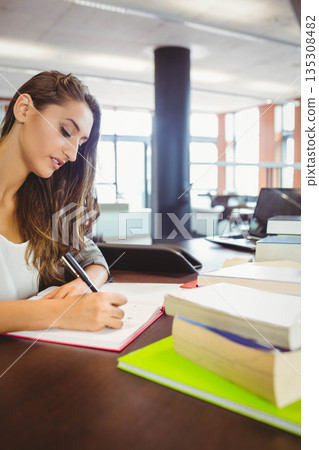 Teenage woman sitting at wooden table in study hall writing in red notebook with stacked textbooks 135308482