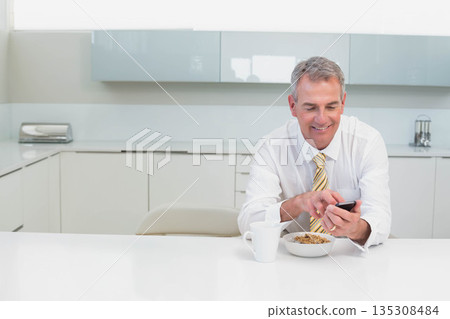 Senior man in business attire holding smartphone while eating cereal at modern kitchen, copy space 135308484