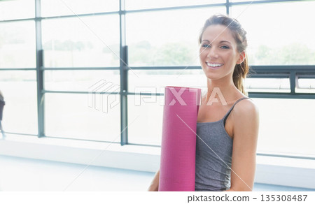 Woman in workout clothes holding pink yoga mat, smiling near windows in fitness studio, copy space Woman in workout clothes holding pink yoga mat, smiling near windows in fitness studio, copy space 135308487
