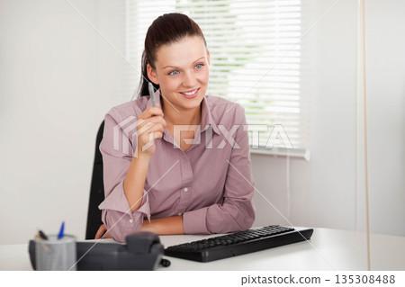 Woman sitting at office desk holding pen, talking while smiling by keyboard and pen holder 135308488