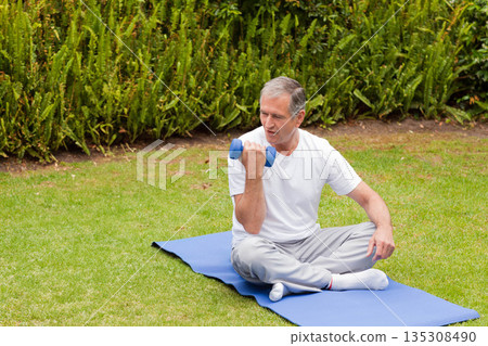 Senior man performing bicep curl on blue exercise mat on grass lawn with blue dumbbell 135308490