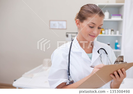Female healthcare professional writing on clipboard with stethoscope in exam room beside exam table Female healthcare professional writing on clipboard with stethoscope in exam room beside exam table 135308498