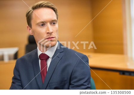 Man in his thirties sitting behind wooden desk in courtroom wearing navy suit and red tie 135308500