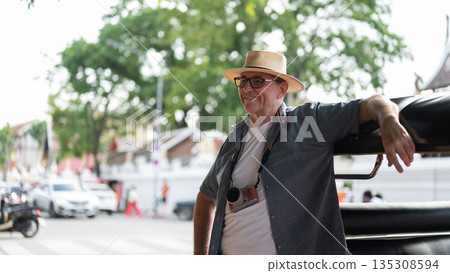 Old white man tourist standing leaning against a tuk tuk or three wheeled taxi after touring a city. 135308594