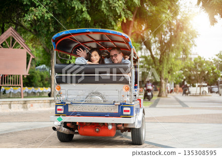 Old white man and asian woman waving a hand while riding in tuk tuk three wheel taxi touring a city. 135308595
