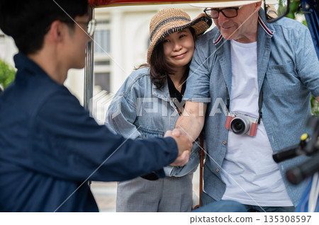 Old white man handshaking a driver aside wife after riding in a tuk tuk or three wheeled taxi touring a city. 135308597