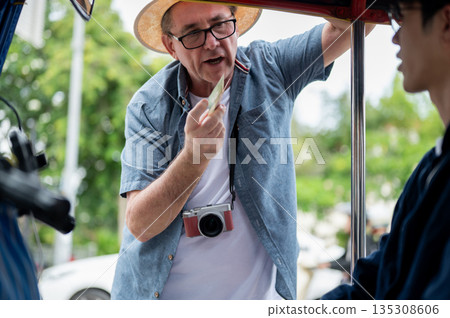 Old white man paying fare to asian driver after riding in tuk tuk or three wheel taxi touring a city 135308606