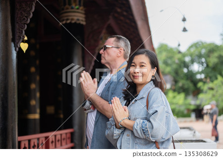 Old white man and smiling asian woman joining palms praying worshipping at ancient buddhist temple. Old white man and smiling asian woman joining palms praying worshipping at ancient buddhist temple. 135308629
