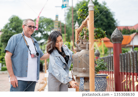 Old white man looking at asian woman ringing a brass bell for luck and prosperity at buddhist temple 135308630