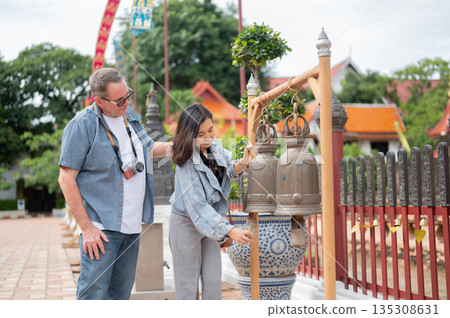 Old white man looking at asian woman ring the brass bell for luck and prosperity at buddhist temple. 135308631
