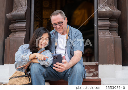 Old white man holding phone looking at photos taken with a woman on wooden steps of buddhist temple. Old white man holding phone looking at photos taken with a woman on wooden steps of buddhist temple. 135308640