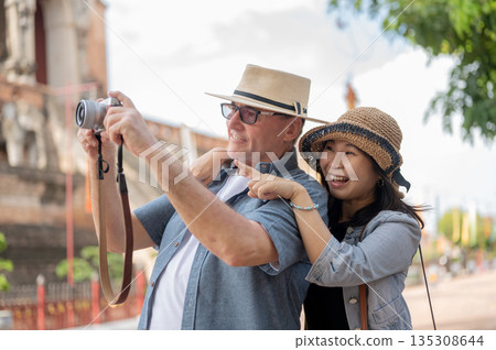Old white man holding camera taking pictures hugged by asian woman standing in a buddhist temple grounds. 135308644