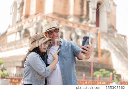 Old white man holding phone taking picture with asian woman standing aside ancient pagoda in temple. 135308662