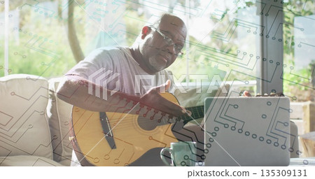 Sitting senior man wearing glasses holding guitar on sofa in studio, with laptop and coffee mug Sitting senior man wearing glasses holding guitar on sofa in studio, with laptop and coffee mug 135309131