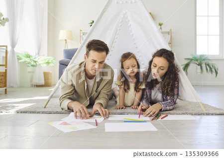 Happy parents and daughter drawing together on floor at home in cozy play tent Happy parents and daughter drawing together on floor at home in cozy play tent 135309366