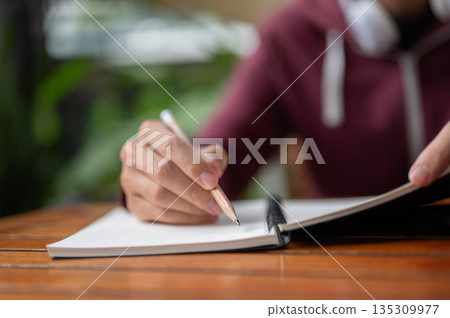 Close up college student man holding pencil writing or drawing in notebook at wooden table in a cafe Close up college student man holding pencil writing or drawing in notebook at wooden table in a cafe 135309977