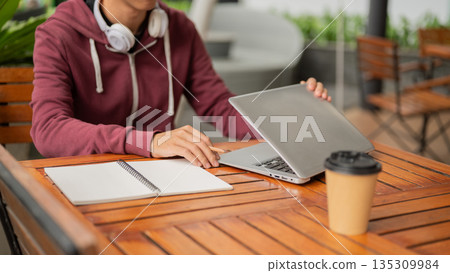 A man or student holding laptop lid closing or opening while sits with coffee at wood table in cafe. 135309984