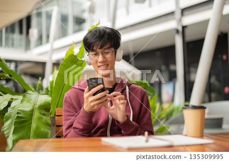 Glasses student man wears headphone smiling while looking at phone sits with notebook at cafe table. 135309995