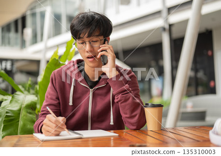 Glasses student man talking on phone writing in notebook while sitting at wooden table outside cafe. 135310003