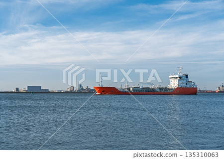 Orange tanker ship sailing on sunny day with blue sky and cityscape on the horizon. 135310063