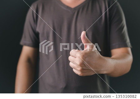 A person in a casual dark shirt gives a thumbs-up gesture against a black background, symbolizing approval, agreement, positivity, encouragement, satisfaction, or successful outcomes. 135310082