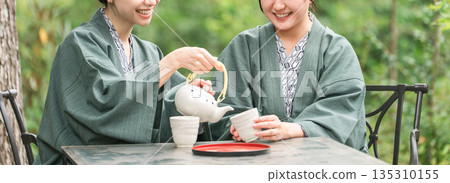 一位年輕的亞洲女子身穿浴衣,在旅途中一邊喝茶一邊享受森林浴。 一位年輕的亞洲女子身穿浴衣,在旅途中一邊喝茶一邊享受森林浴。 135310155