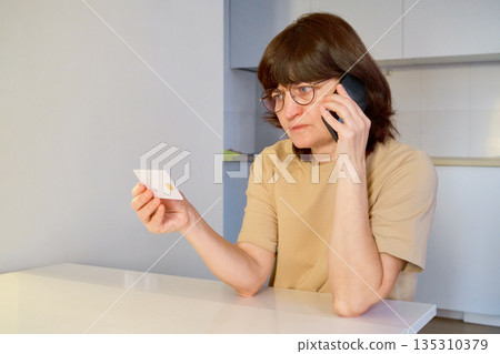 Woman making phone call, holding credit card and showing worried expression 135310379
