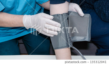 Close-up of a healthcare professional's hands in white medical gloves applying a blood pressure cuff to a patient's arm. Concept of hypertension control, medical check-up, and elderly care at home or Close-up of a healthcare professional's hands in white medical gloves applying a blood pressure cuff to a patient's arm. Concept of hypertension control, medical check-up, and elderly care at home or 135312041