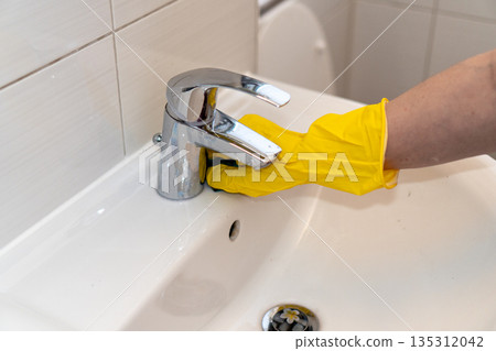Close-up of a person in yellow protective rubber gloves wiping a chrome water tap on a white ceramic sink. Concept of professional cleaning service, home hygiene, and disinfection Close-up of a person in yellow protective rubber gloves wiping a chrome water tap on a white ceramic sink. Concept of professional cleaning service, home hygiene, and disinfection 135312042