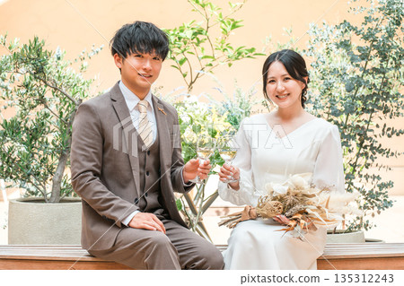 Couple in dresses sitting on chairs drinking champagne, married couple, bride and groom, dressed up couple, millionaire, wealthy 135312243