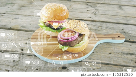Displaying sesame bun burgers on wooden board with blue handle at picnic table, with media icons Displaying sesame bun burgers on wooden board with blue handle at picnic table, with media icons 135312530