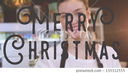Smiling woman in white shirt and dark apron standing behind café counter, with Merry Christmas sign Smiling woman in white shirt and dark apron standing behind café counter, with Merry Christmas sign 135312555