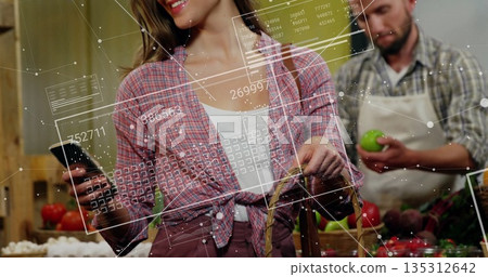 Examining green apples while holding smartphone and basket at grocery stall with digital panels Examining green apples while holding smartphone and basket at grocery stall with digital panels 135312642