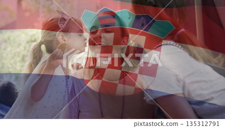 Smiling mom wearing party hat and necklace between daughters snacking in garden under flag overlay 135312791