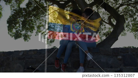 Holding smartphone, two women leaning on stone wall in park under large tree, with bicycle wheel 135312807