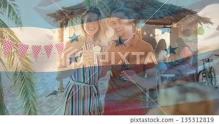Holding cocktail glass and beer bottle, couple standing at beachside tiki bar, with palm trees 135312819