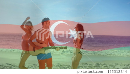 Three friends wearing swimwear playing guitar and dancing at beach water's edge, with flag overlay 135312844