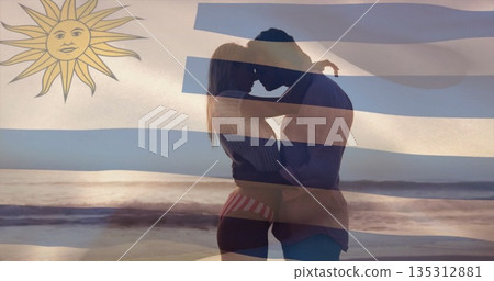Embracing couple in swimwear and shirt hugging on beach at sunset, with Uruguay flag overlay Embracing couple in swimwear and shirt hugging on beach at sunset, with Uruguay flag overlay 135312881