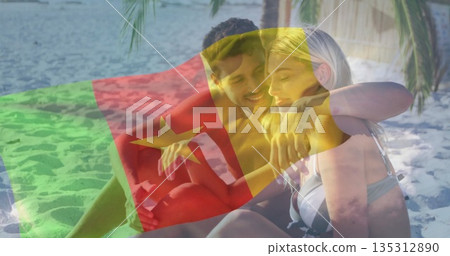 Sitting couple in swimwear embracing each other on sandy beach near calm sea, with flag overlay 135312890