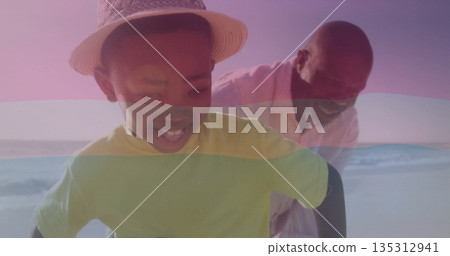 Smiling grandson wearing straw hat playing with grandfather on sandy beach, gentle waves rolling 135312941
