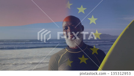 Standing man wearing wetsuit holding yellow surfboard at shore, with EU stars overlay 135312953
