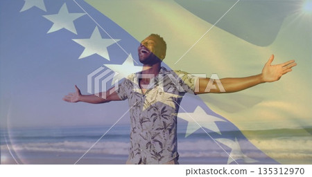 Man wearing tropical shirt raising arms and tilting head back on sandy beach, flag motif overlay 135312970