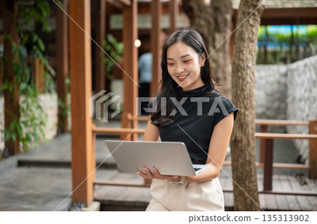 Asian woman or college student holding and looking at laptop while standing in garden courtyard at an outdoor cafe Asian woman or college student holding and looking at laptop while standing in garden courtyard at an outdoor cafe 135313902