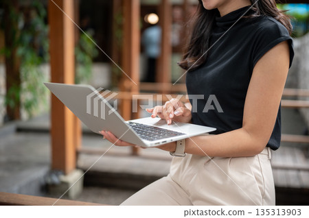 Close up of woman student holding and using laptop while sitting on wooden bench in garden courtyard at outdoor cafe Close up of woman student holding and using laptop while sitting on wooden bench in garden courtyard at outdoor cafe 135313903